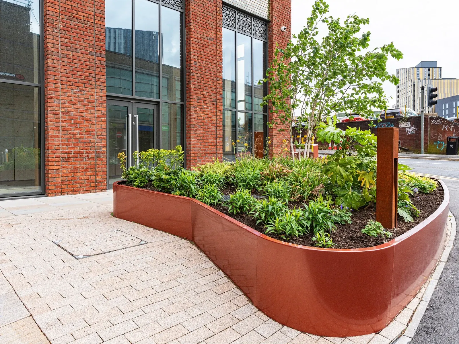 Curved red-brown planter filled with trees and shrubs beside a paved walkway